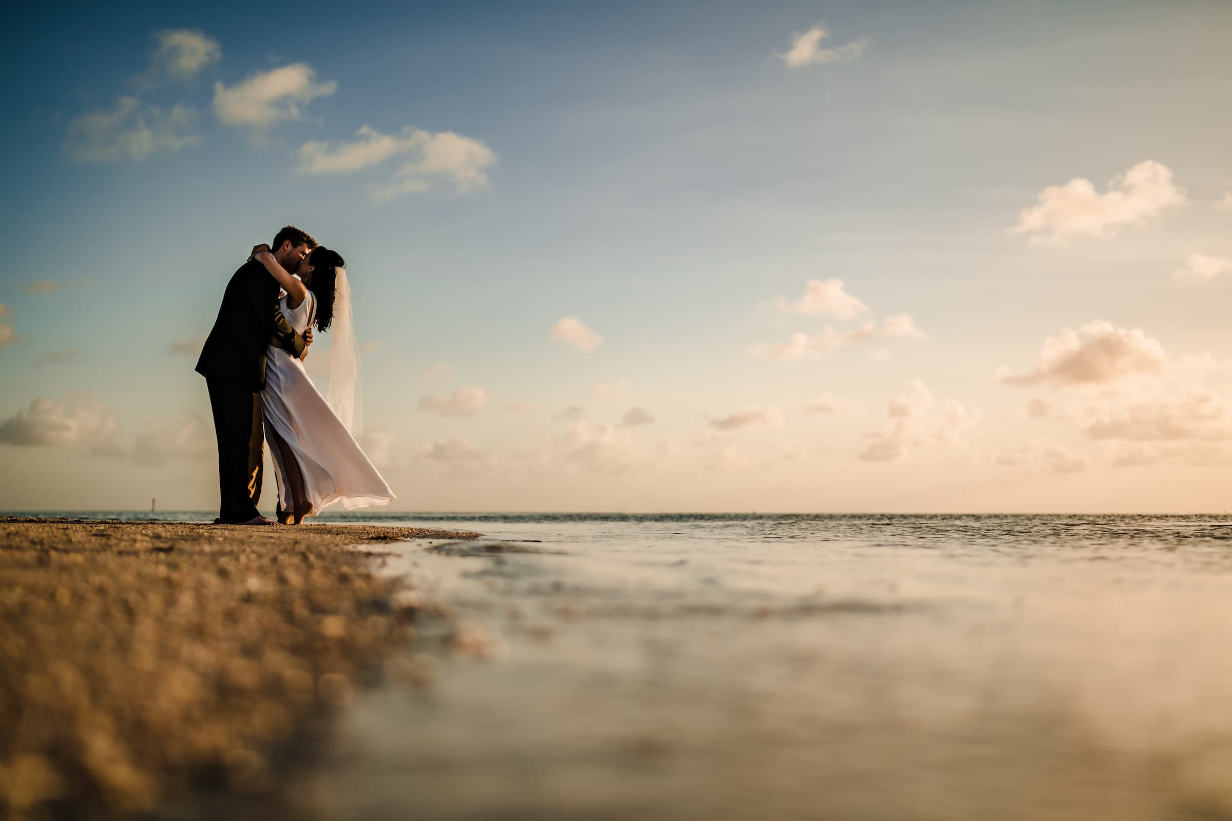 A bride and groom kissing on a beach by the ocean shoreline at Little Palm Island Resort