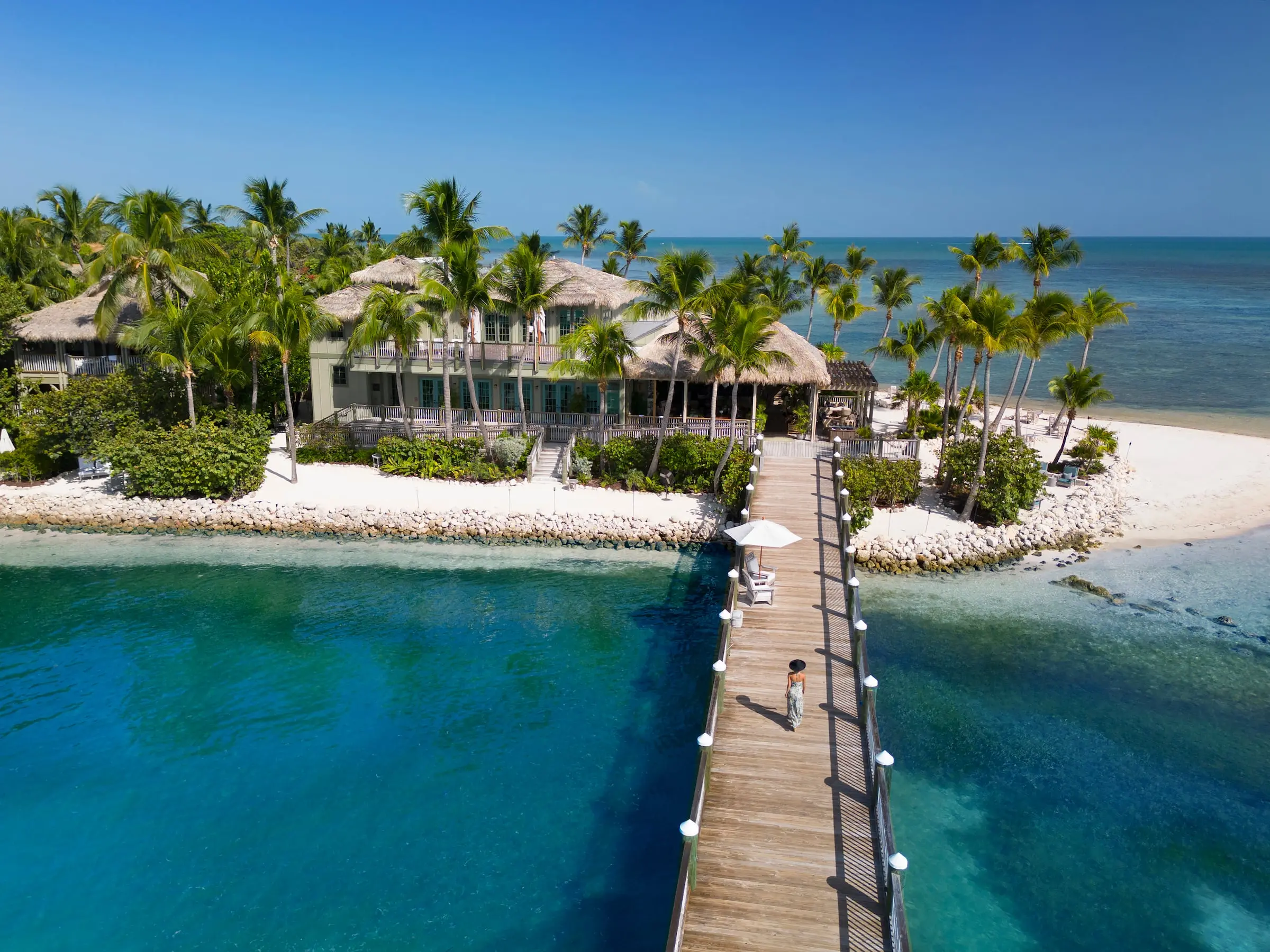 A woman walks along a wooden boardwalk towards the Little Palm Island resort, surrounded by white sand beach